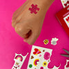 Hand with a temporary tattoo on a pink background with stickers and scissors.