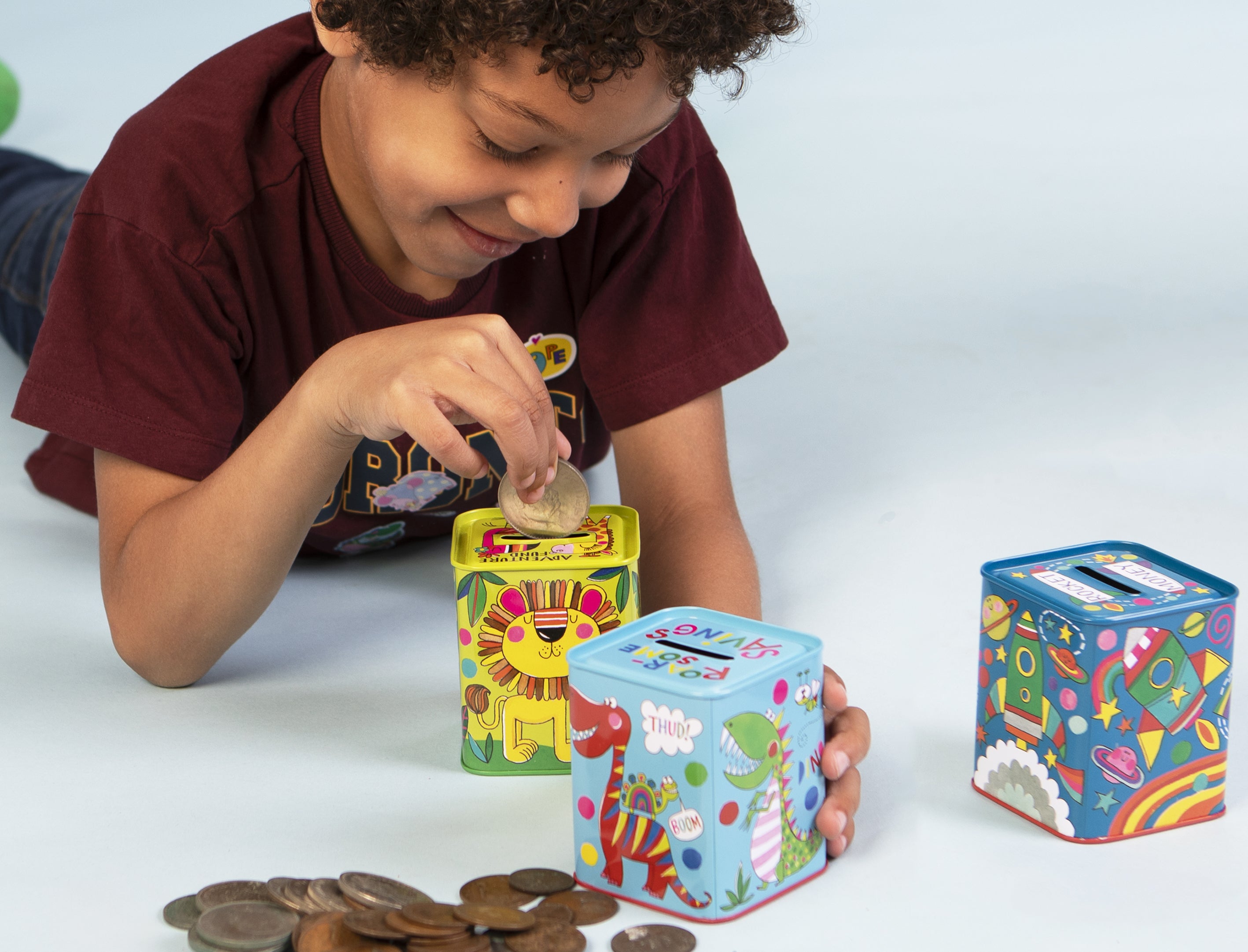 Rachel Ellen Designs children's money boxes or piggy banks; a child is inserting a coin into a lion money tin, with a dinosaur tin nearby.